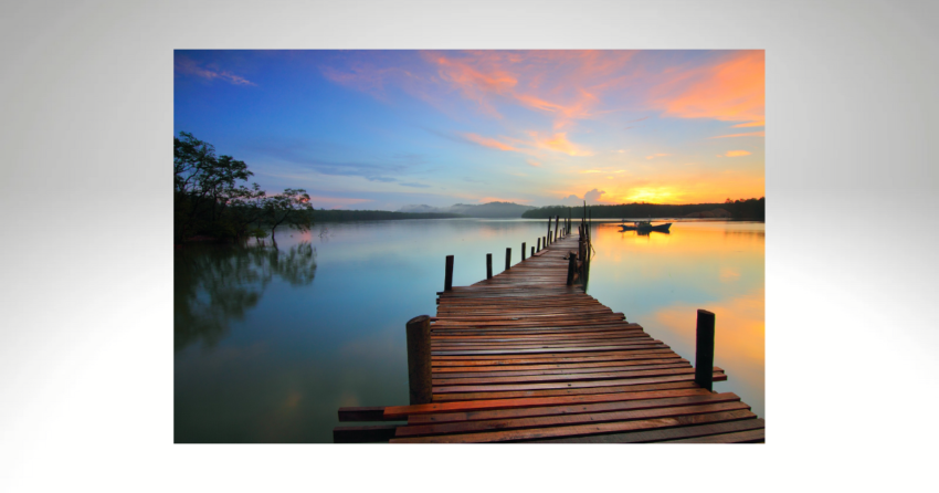 A long wooden dock stretches into a tranquil lake at sunset, reflecting vibrant orange and blue hues. A small boat is anchored in the distance.
